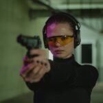 Woman aiming a pistol indoors at a shooting range, wearing eye and hearing protection.